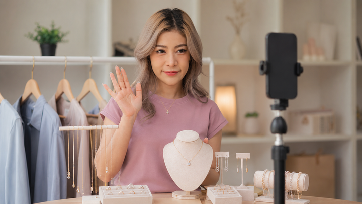 Independent jeweler running a live commerce show from their store using a smartphone tripod, with jewelry displayed on a tray and viewers commenting on a second screen showing live engagement metrics, illustrating live commerce for jewelry in 2026.
